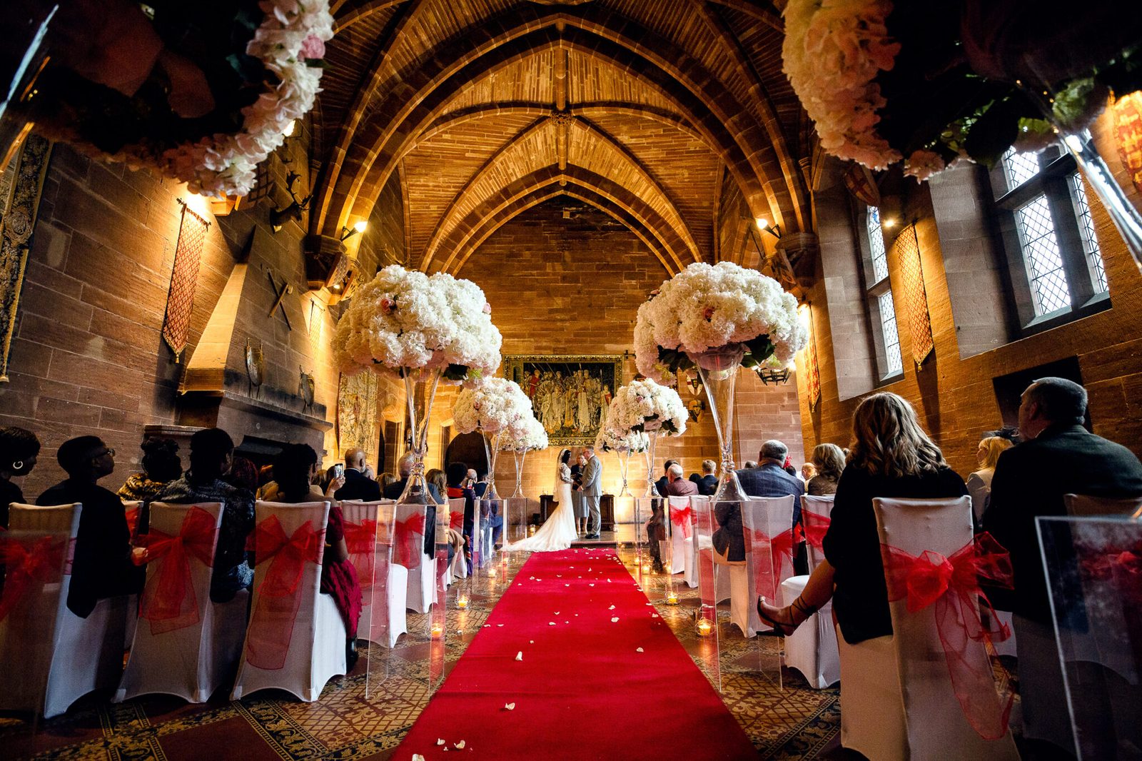 A decorated ceremony room with a red carpet aisle and tall floral arrangements