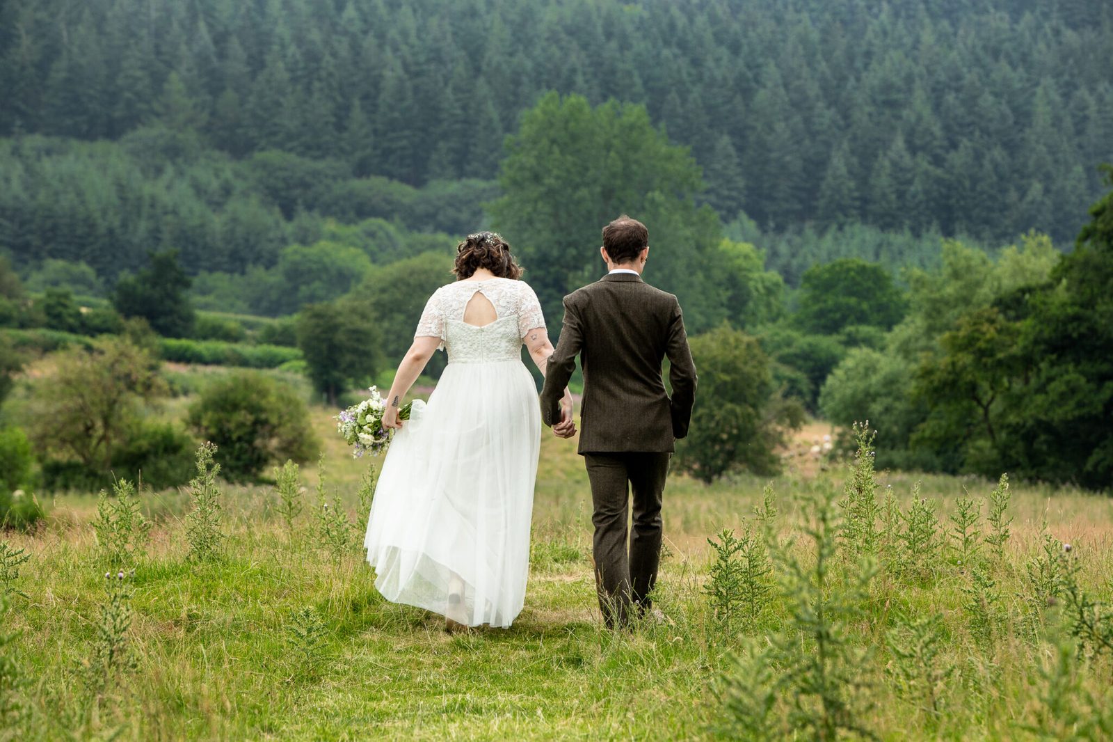 A couple walk away through a long-grass meadow towards wooded hills