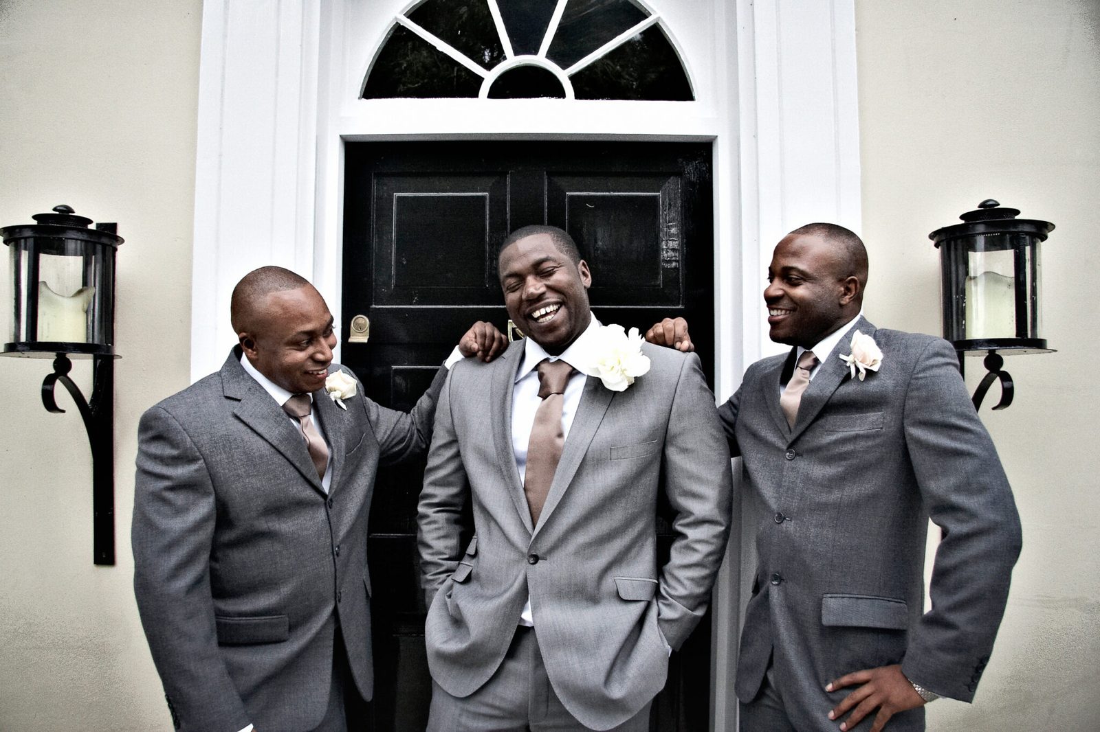A groom laughs with two groomsmen in grey suits outside a black front door