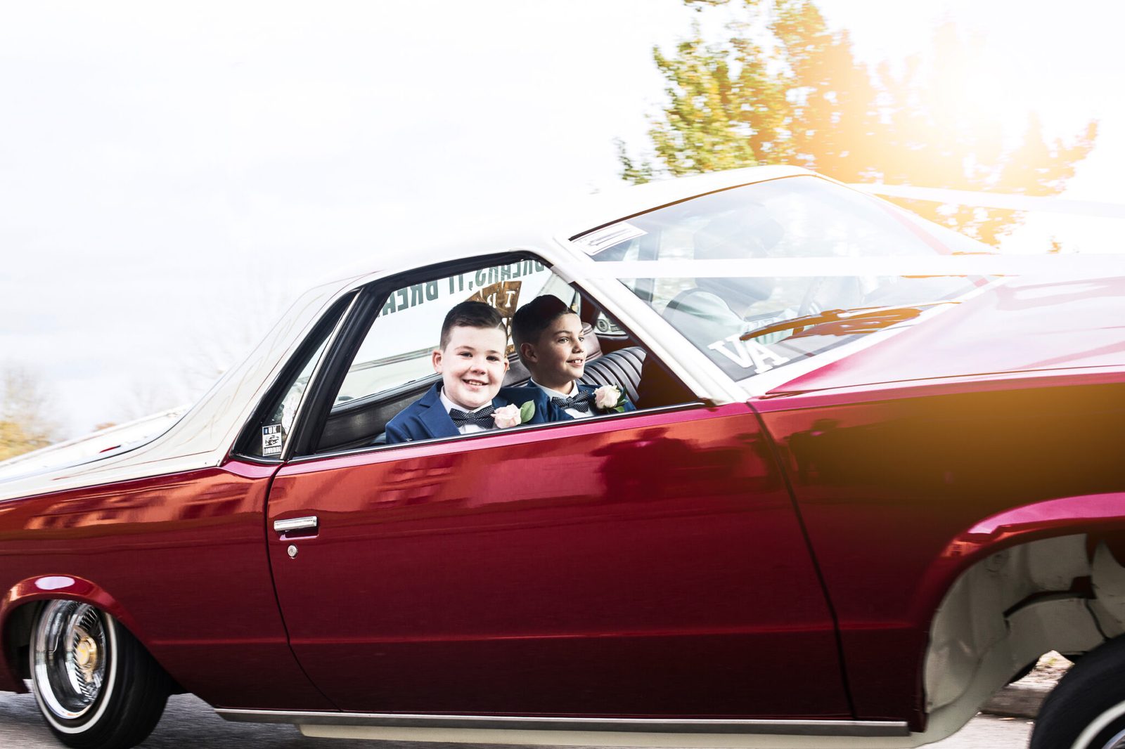 Two boys grin from the window of a red classic car