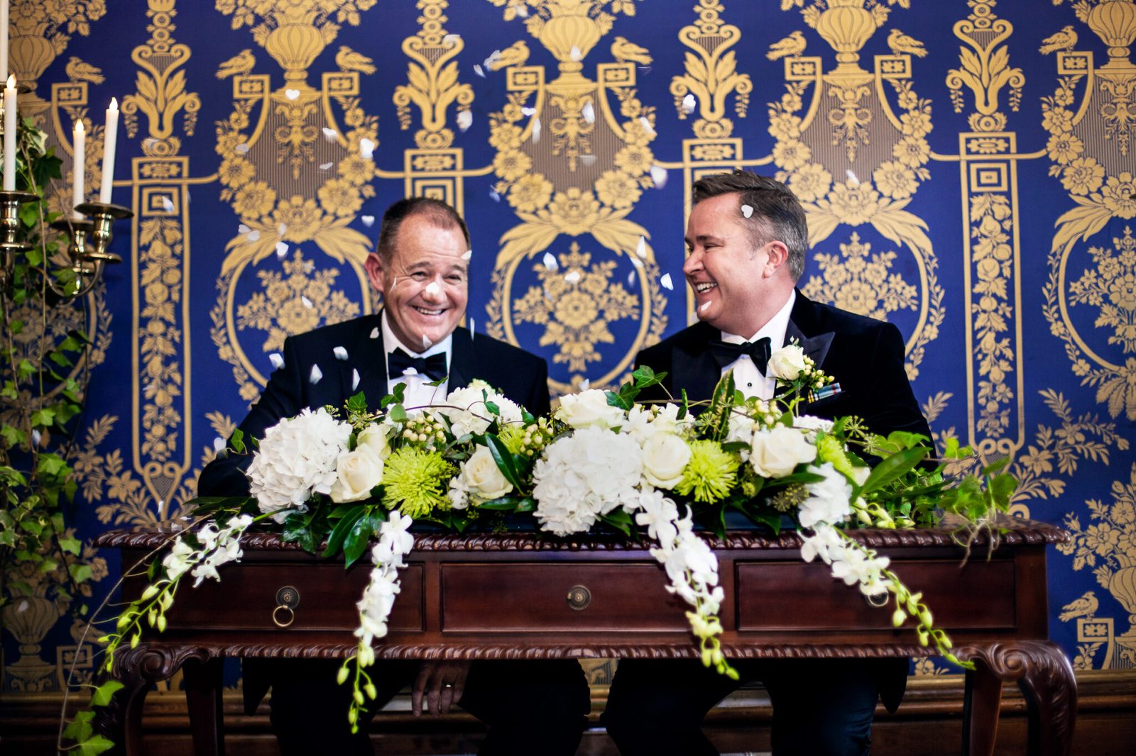 Two grooms smile as they sign the register before a blue patterned wall
