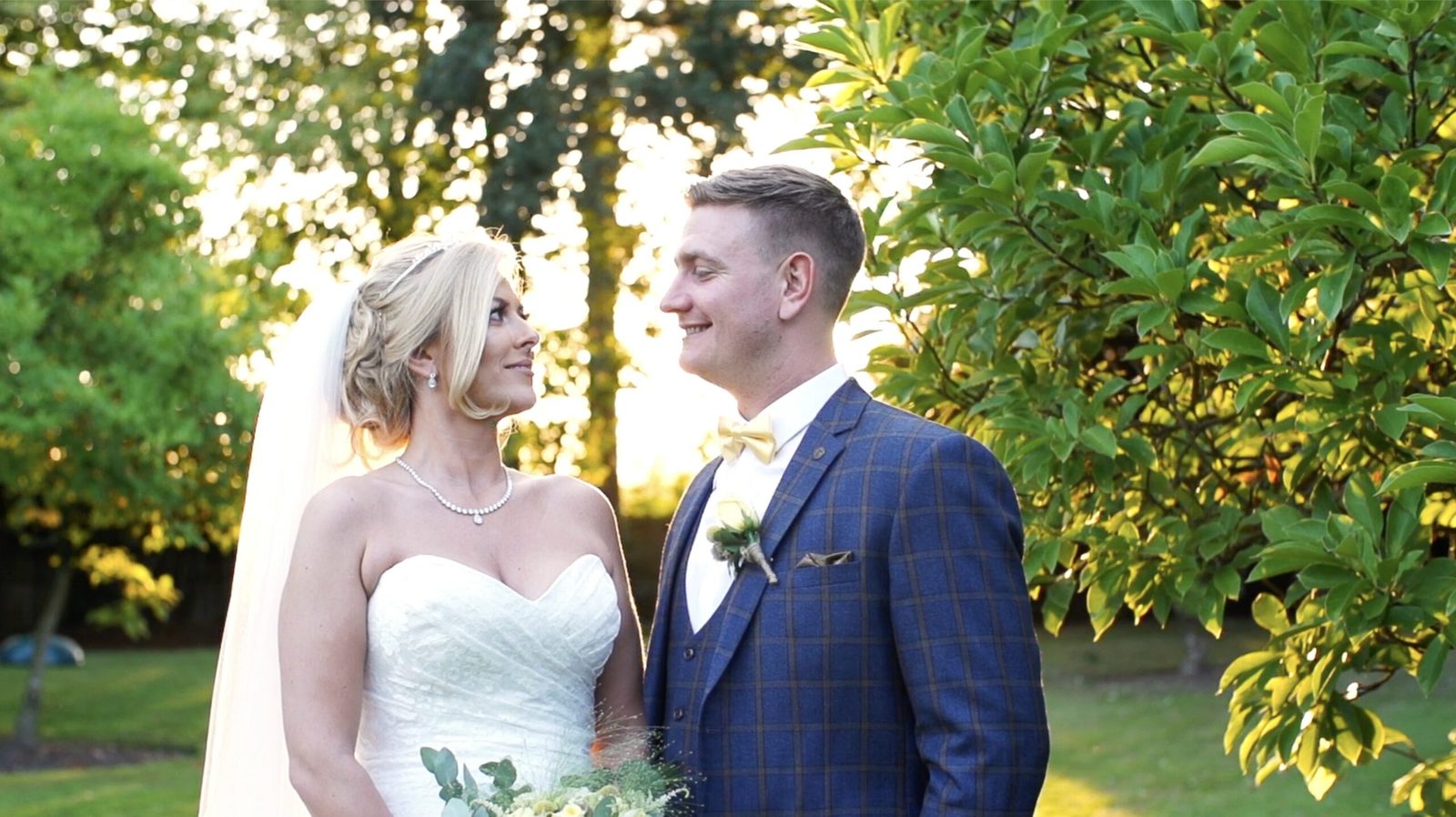 A bride and groom smile at each other beneath sunlit trees — a still from the wedding film