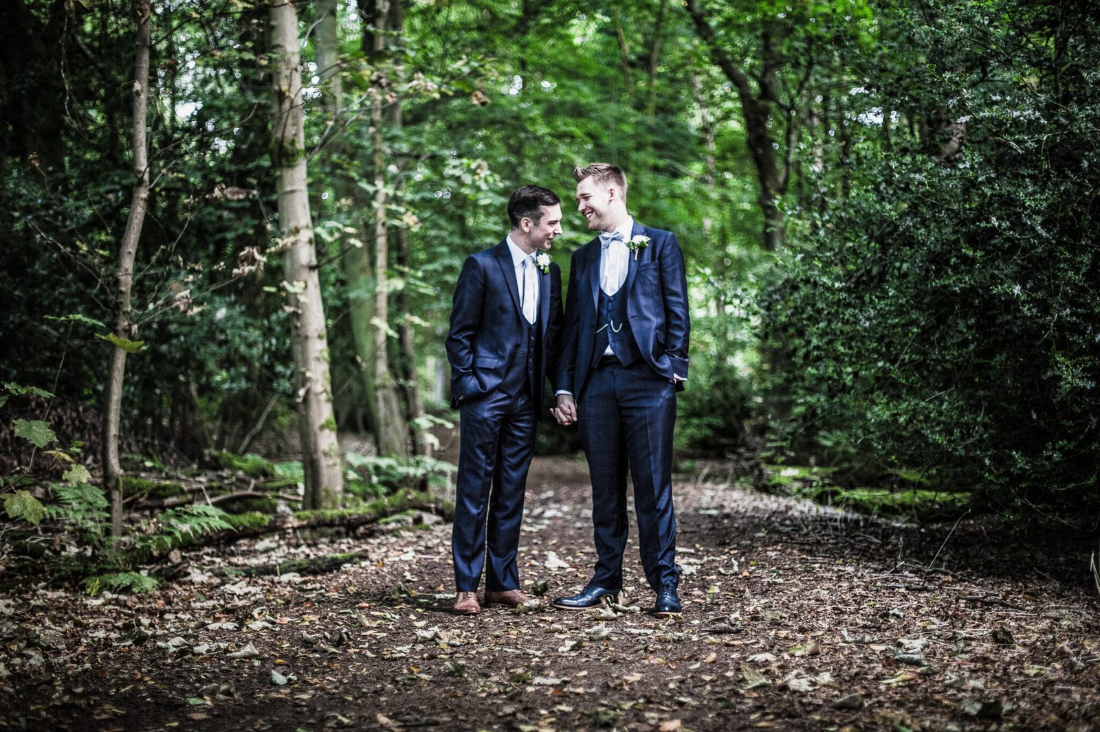 Two grooms stand together on a woodland path