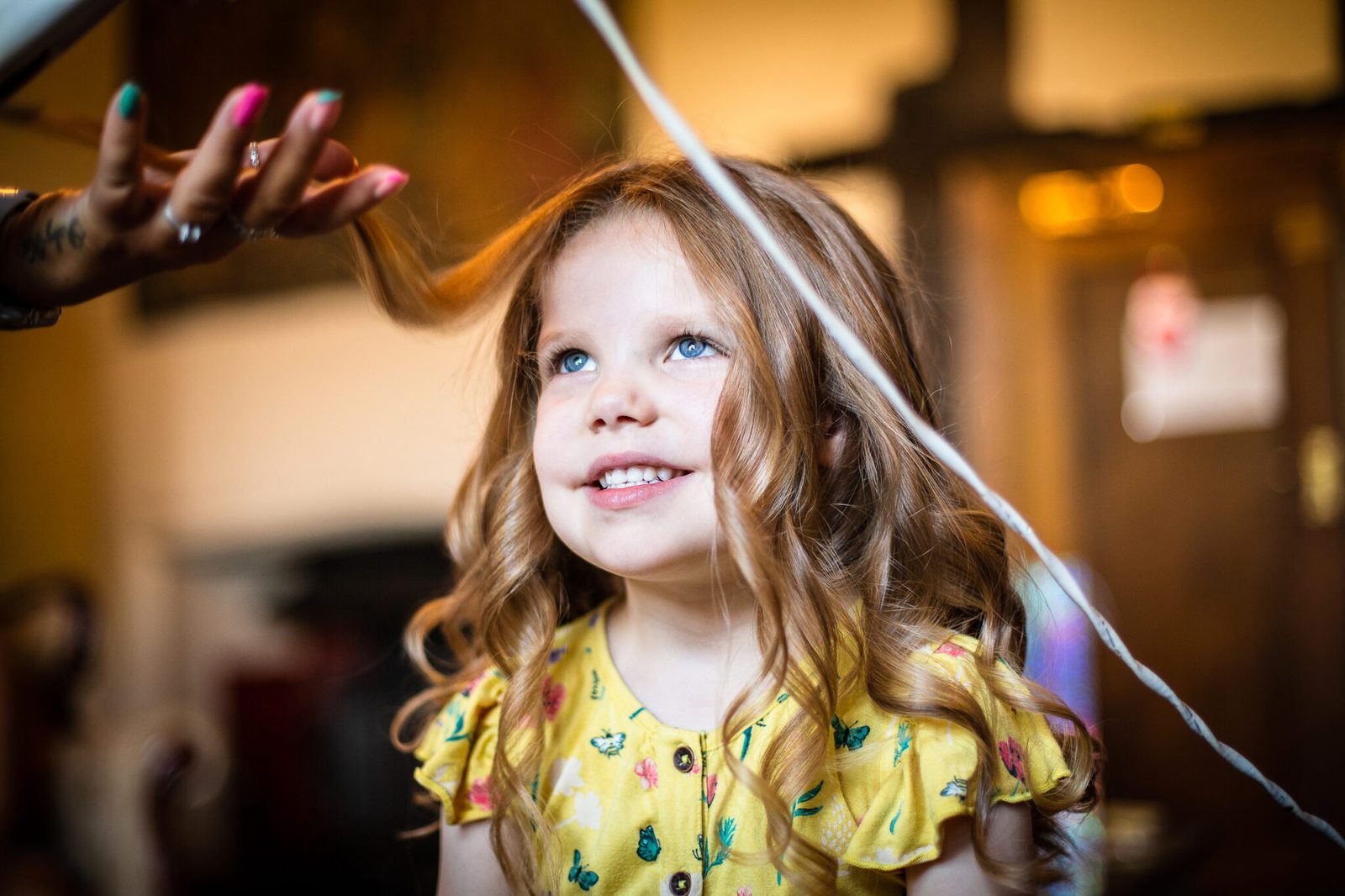 A little girl in a yellow dress looks up while her hair is styled