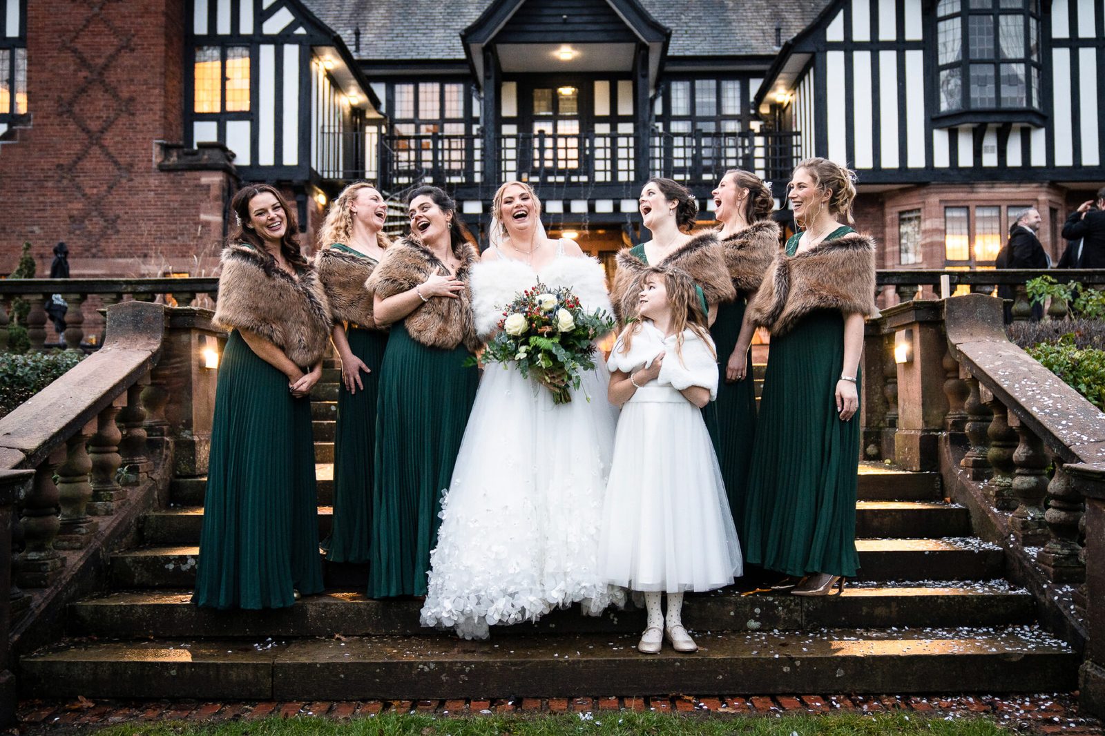 A bride with bridesmaids in green on the steps of a timber-framed hall