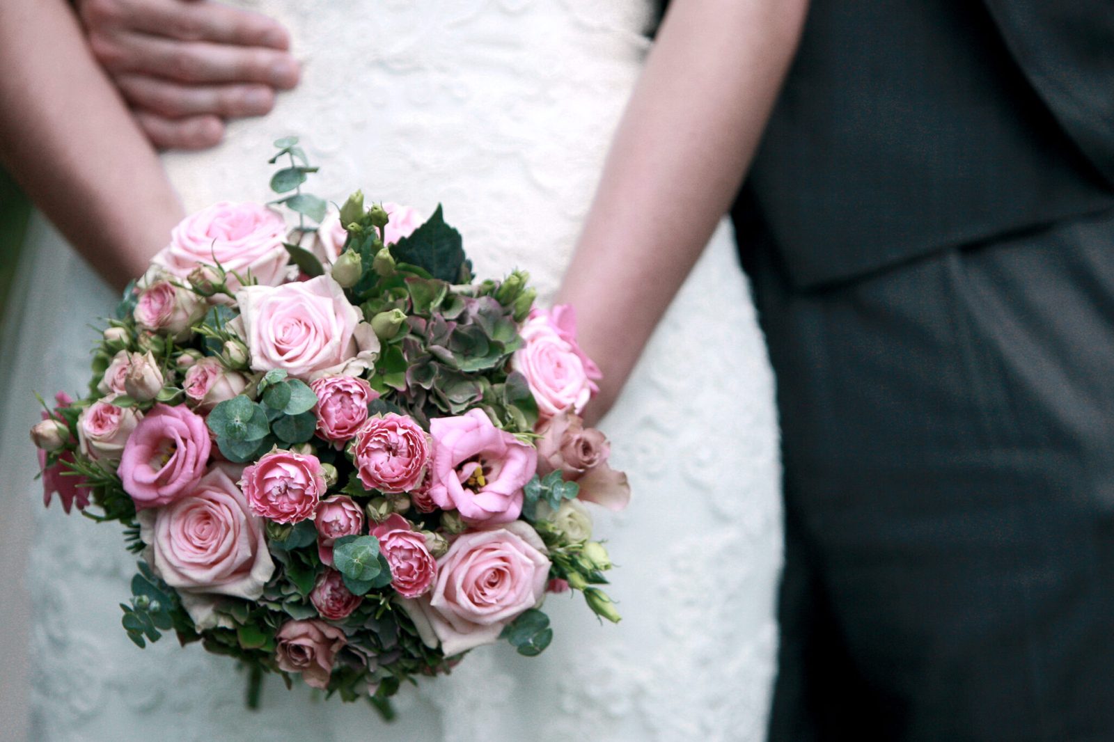 A bride holds a pink rose bouquet beside the groom's dark suit