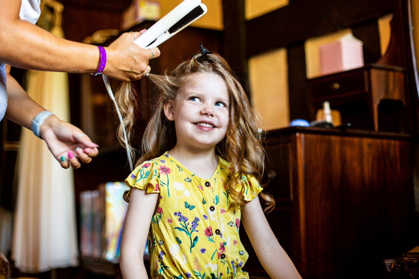 A little girl in a yellow dress smiles while her hair is curled
