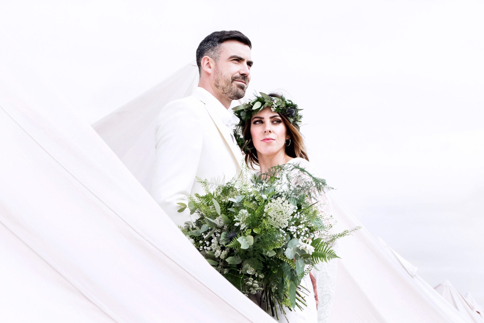 A couple dressed in white with a greenery bouquet against a pale sky