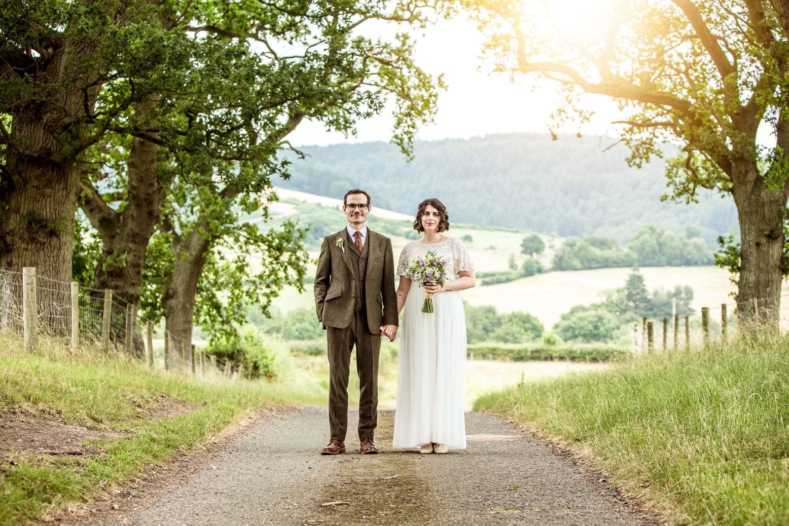 A couple stand on a country lane with rolling hills behind