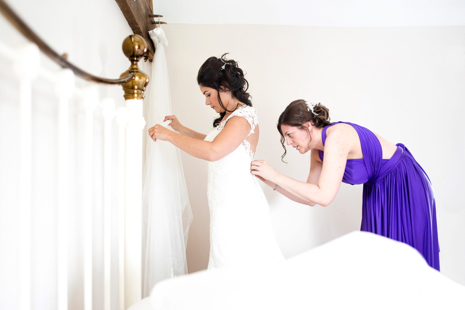 A bridesmaid in purple fastens the back of the bride's dress