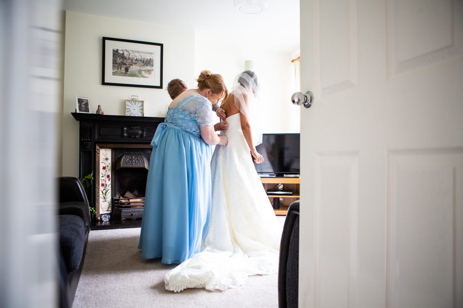 A bridesmaid in blue fastens the bride's dress in a living room