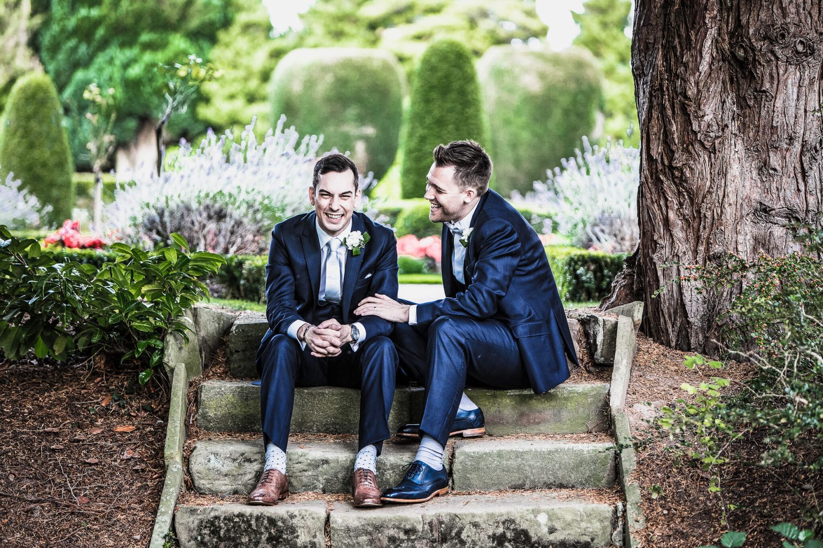 Two grooms sit together on stone steps in a topiary garden