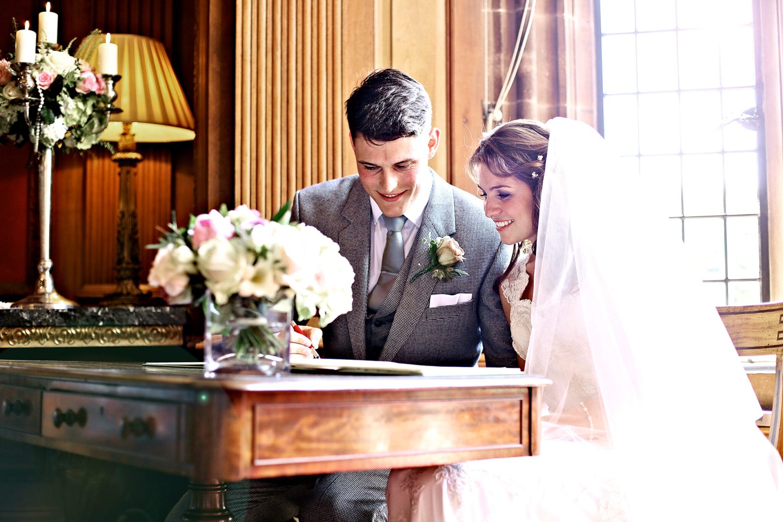 A groom and bride smile as they sign the register at a wooden desk
