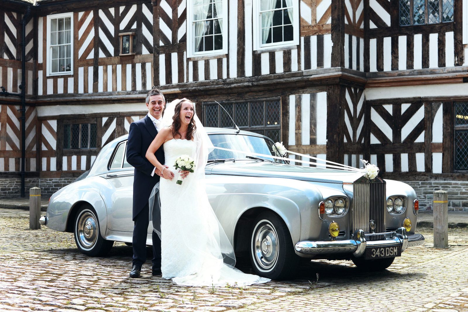 A couple beside a silver vintage Rolls-Royce outside a black-and-white timber-framed hall