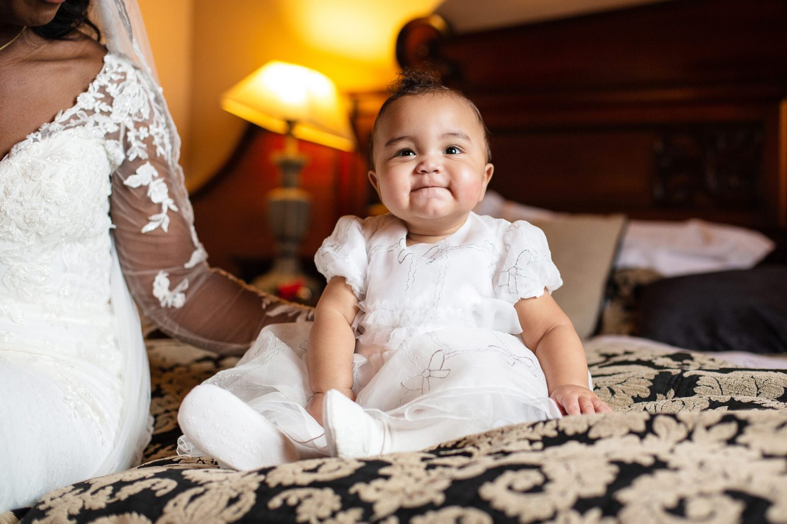 A baby in a white outfit sits up on a patterned bedspread