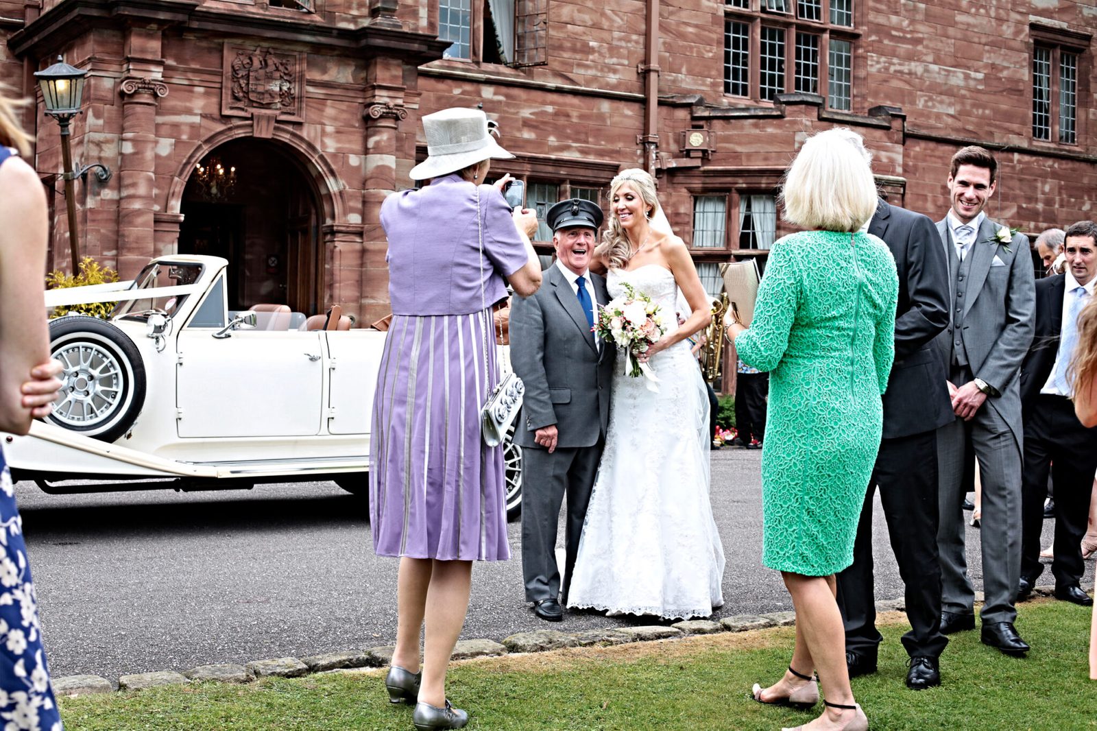 The bride and guests outside a red-brick building beside a vintage car