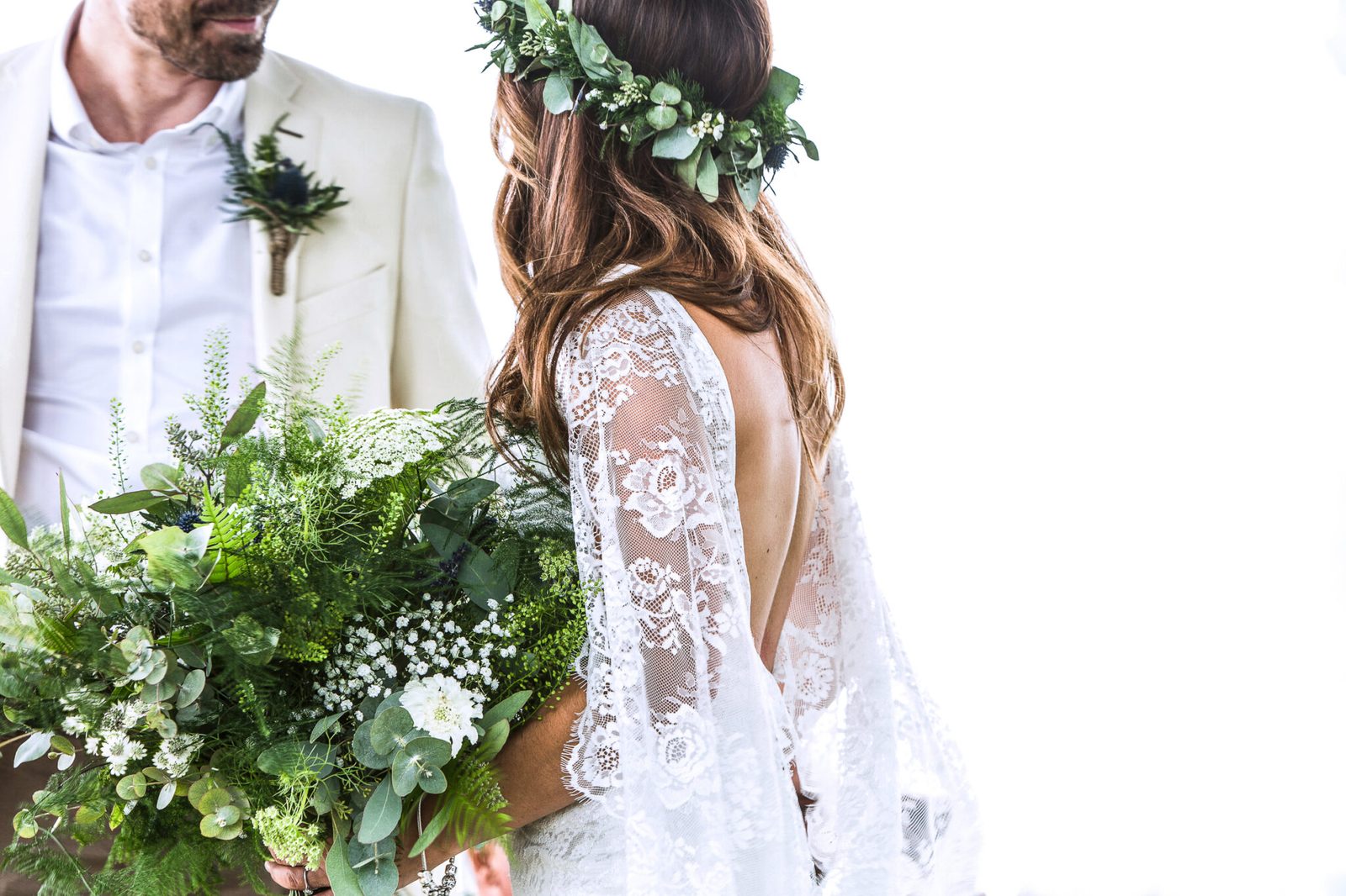 A bride in a flower crown and lace dress beside the groom, greenery bouquet in hand