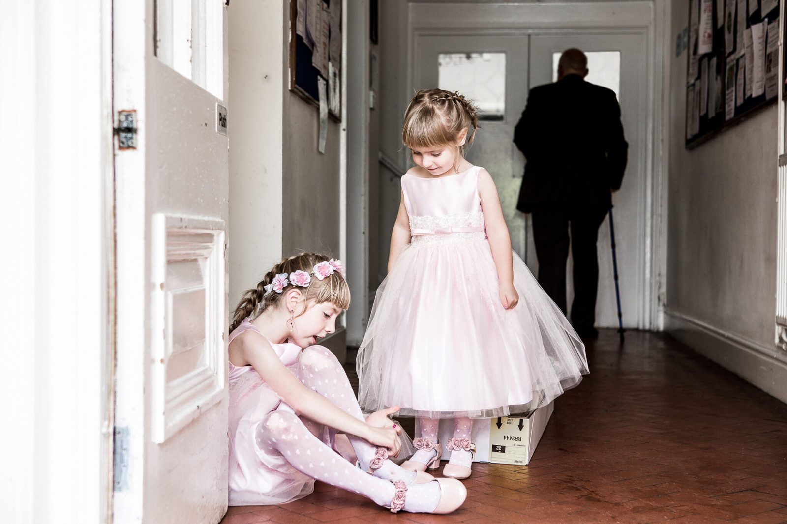 A flower girl in a pink dress waits in a hallway while another child sits behind her