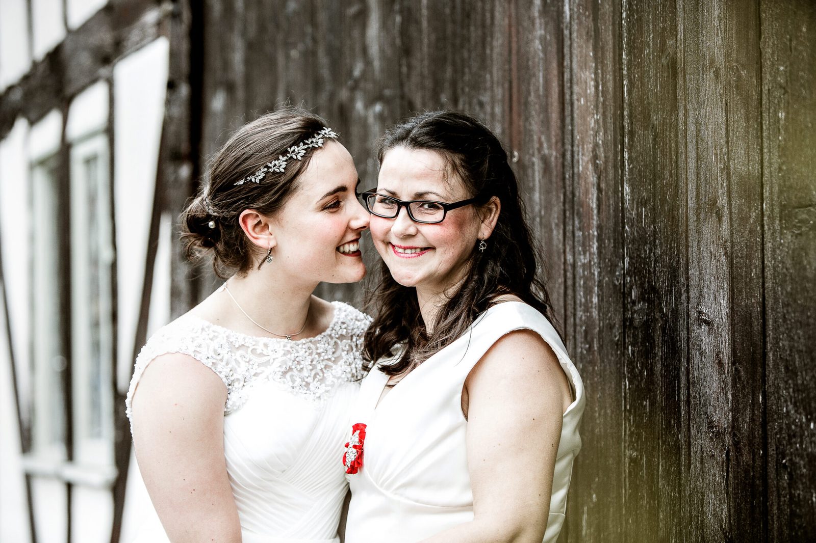 Two brides laugh close together beside a timber wall