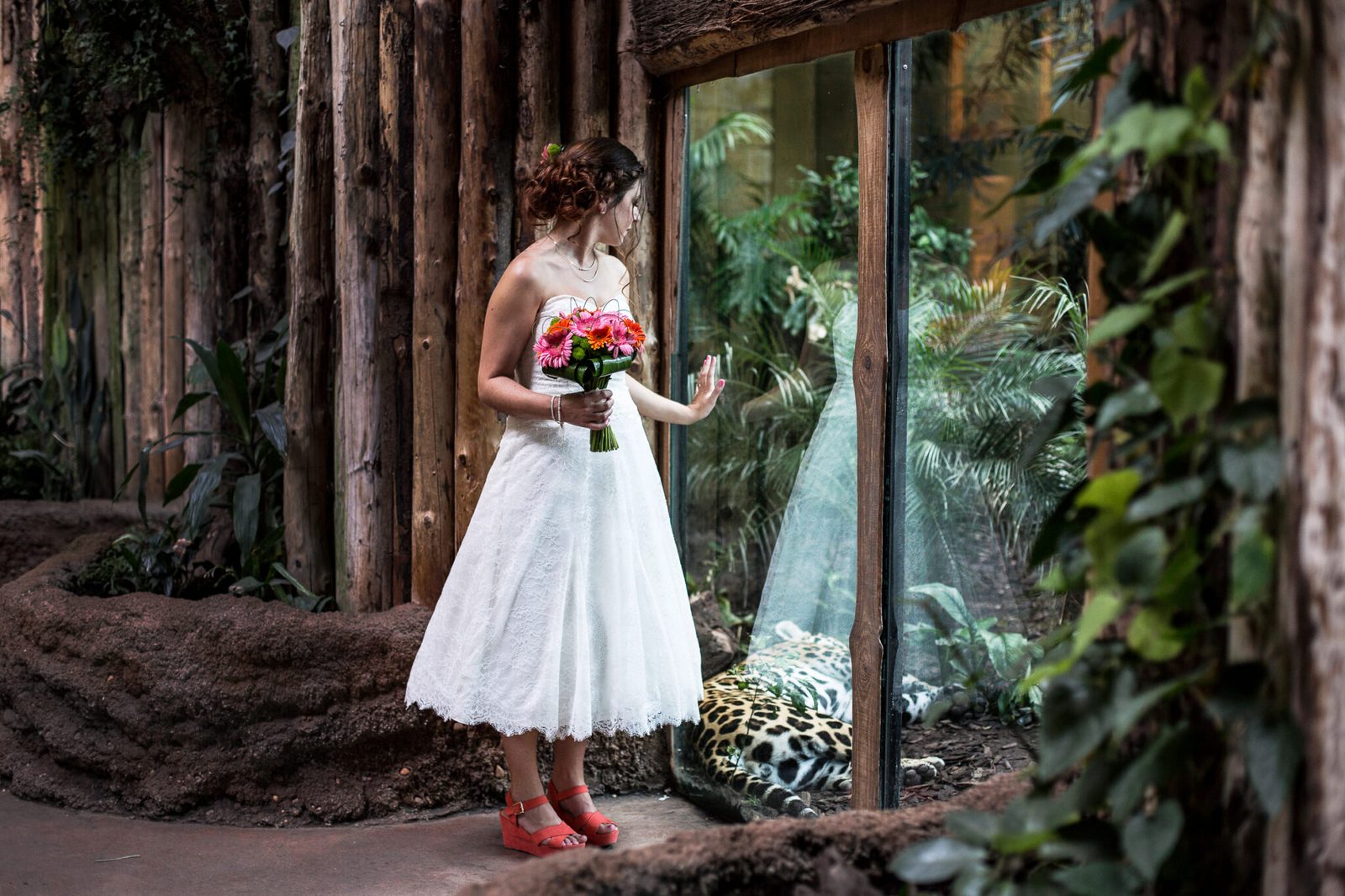 A bride holding a bouquet stands by a glasshouse window